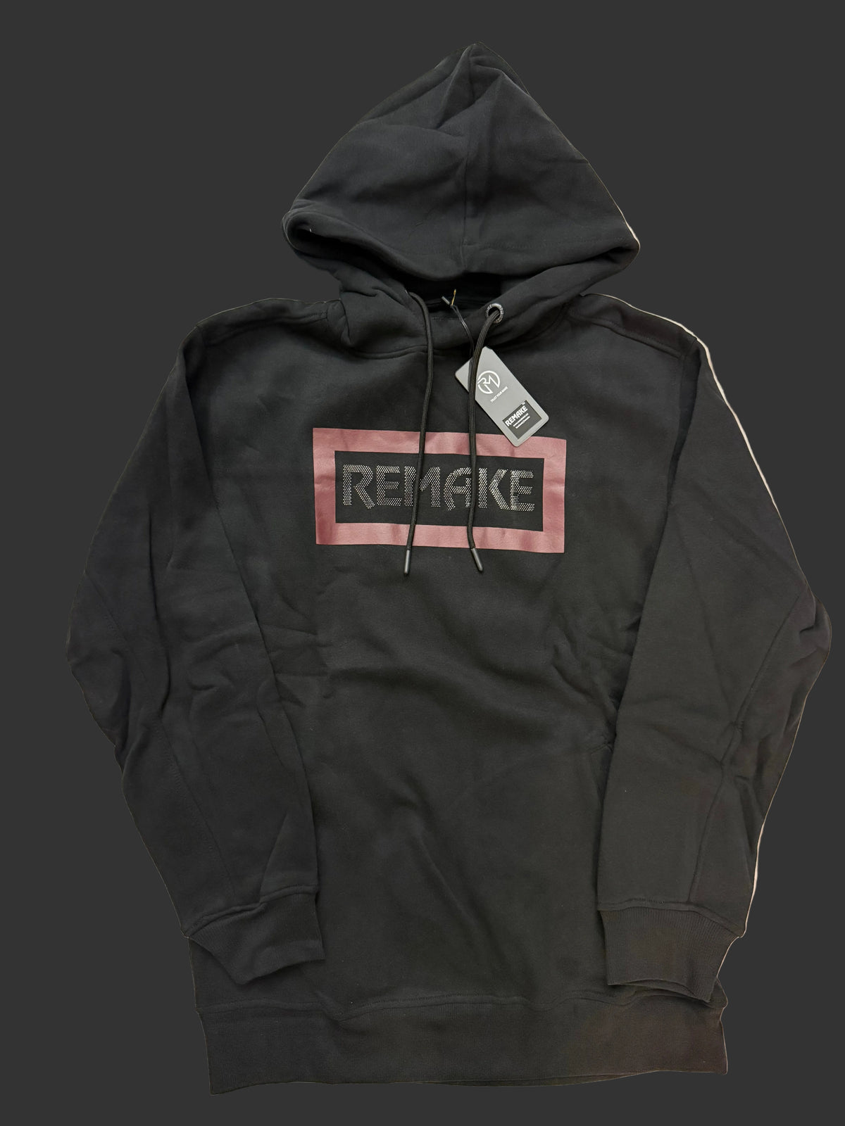 A black pullover hoodie with a graphic chest print reading 'REMAKE' inside a dusty pink rectangular border, laid flat on a light beige fabric background. A hangtag is attached near the collar.