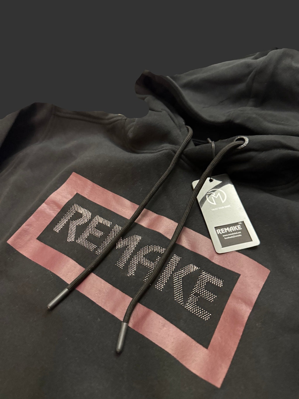 A black pullover hoodie with a large graphic print on the front. The graphic features a dusty pink rectangular border surrounding a black center block with the word 'REMAKE' in a textured, metallic silver font.