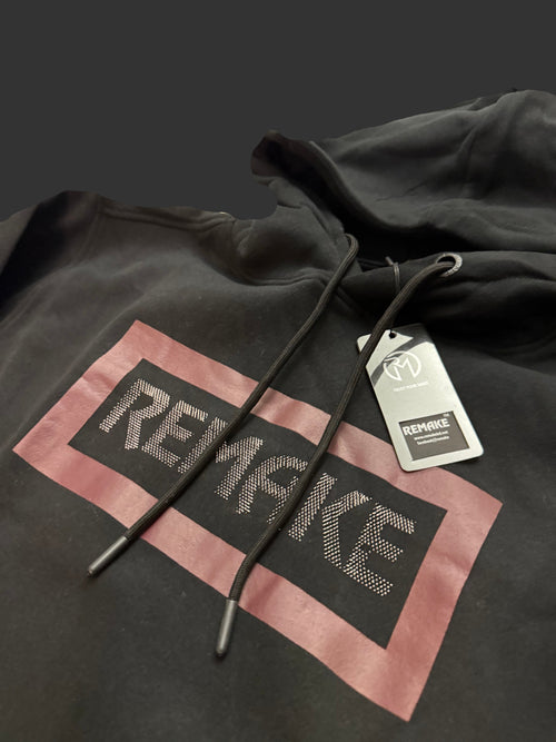A black pullover hoodie with a large graphic print on the front. The graphic features a dusty pink rectangular border surrounding a black center block with the word 'REMAKE' in a textured, metallic silver font.