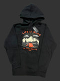 A black pullover hoodie laid flat on a light fabric background, featuring a front graphic with the text 'LIFE IS GOOD' and 'IN THE WOODS' around an illustrated mountain scene with a red tent.