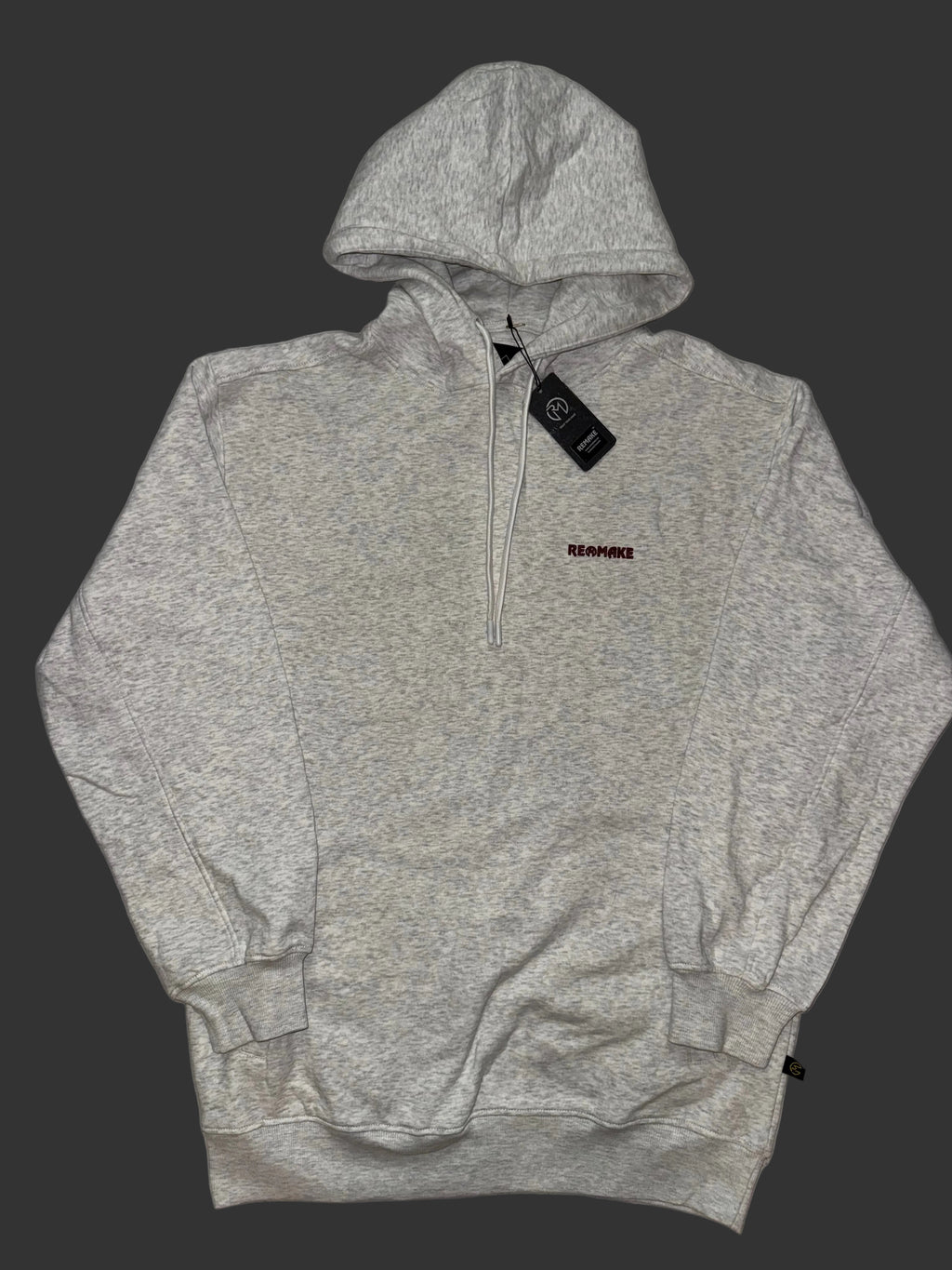 A oatmeal gray pullover hoodie with a half-zip neckline, featuring a small red 'REMAKE' logo on the left chest and a black hangtag, laid flat on a light gray patterned fabric background.
