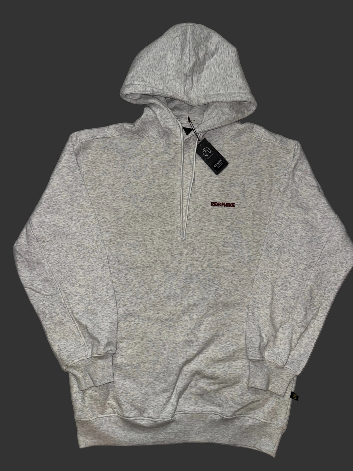A oatmeal gray pullover hoodie with a half-zip neckline, featuring a small red 'REMAKE' logo on the left chest and a black hangtag, laid flat on a light gray patterned fabric background.